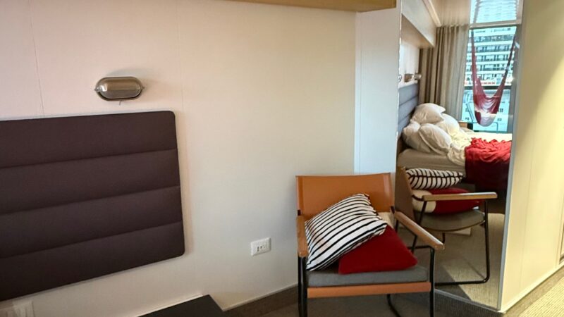 Virgin Voyages cruise ship cabin featuring a chair with pillows, a cushioned wall panel, and a glimpse of a bed with a hammock by the window.