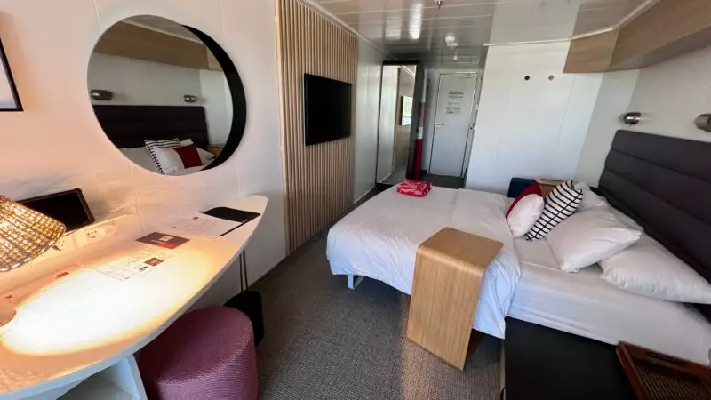 Virgin Voyages cruise ship cabin with a neatly made bed, pillows, a small table, a wall mirror, and desk with a lamp and documents.