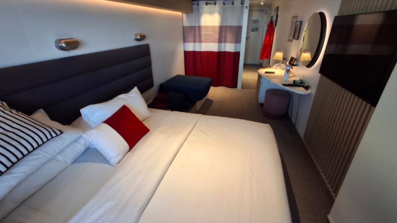Elegant Virgin Voyages cruise ship cabin with a large bed, modern decor, wall-mounted TV, and a workspace with a round mirror.