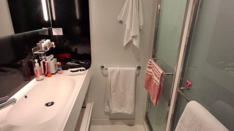 Modern Virgin Voyages cruise ship cabin bathroom with a sleek sink, large mirror, glass shower door, and neatly hung towels.