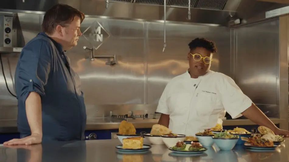 Two chefs in a stainless-steel kitchen with plated dishes on a counter