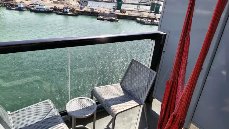 Cruise ship balcony with two metal chairs, a small table, and a red hammock, overlooking the harbor.