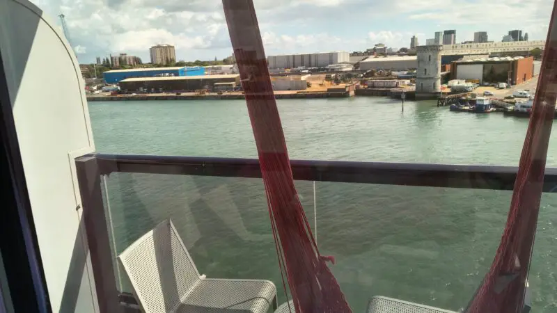 Balcony view from a Virgin Voyages cruise ship cabin, featuring a chair and red hammock with a waterfront and buildings in the background.