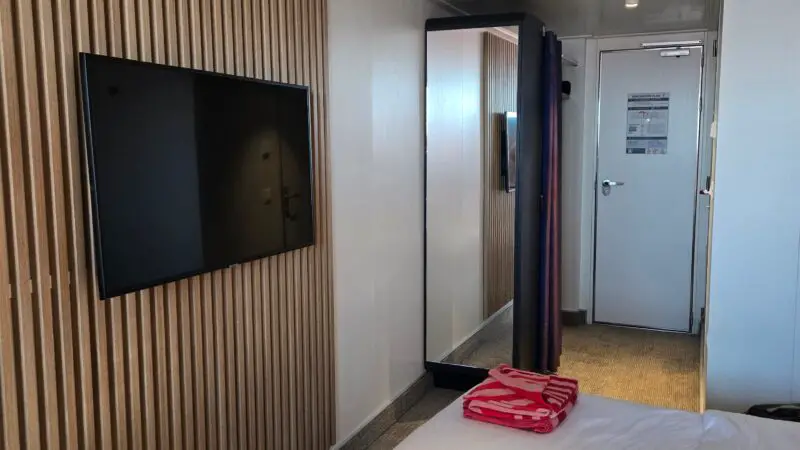 Cruise ship cabin with wood-paneled wall, a flat-screen TV, a bed with white linens and a pink towel, a full-length mirror, and a closed door.