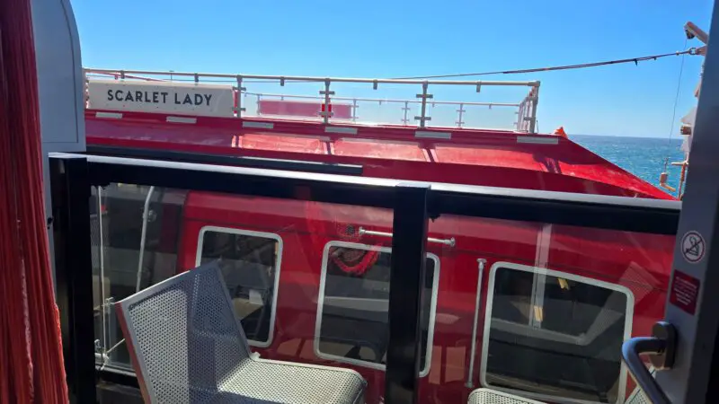 View from a Virgin Voyages balcony cabin showing a bright red ship structure labeled "Scarlet Lady" outside, with ocean and sky in the background. The balcony has two perforated chairs and a small table.