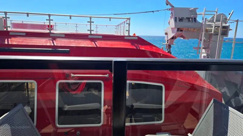 View from a Virgin Voyages cruise ship balcony cabin, featuring glass railing, two chairs, and an ocean view with a lifeboat and the sea visible beyond.