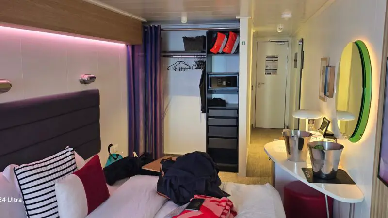 Virgin Voyages cruise ship cabin with a bed, modern decor, wall-mounted lights, open closet, and vanity area with a round mirror.