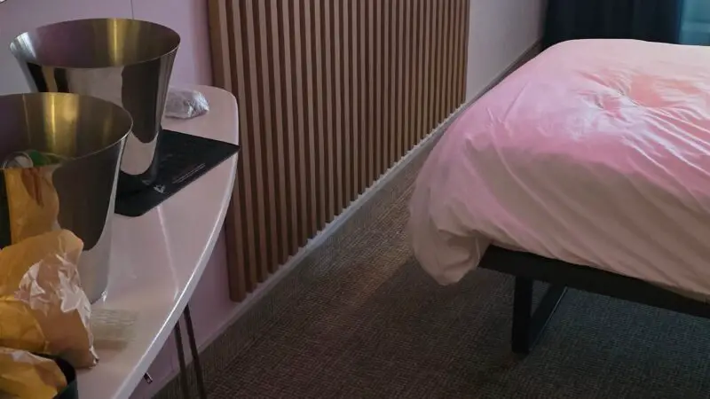 Virgin Voyages cruise ship cabin interior with a modern design, featuring a bed, wall-mounted TV, round mirror, and a table with items on it.