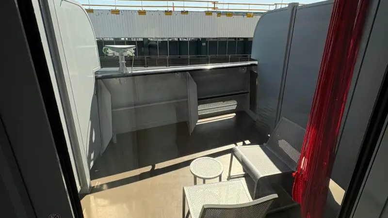View of a Virgin Voyages cruise ship balcony with two chairs, a table, and a red hammock. The cabin overlooks the port.