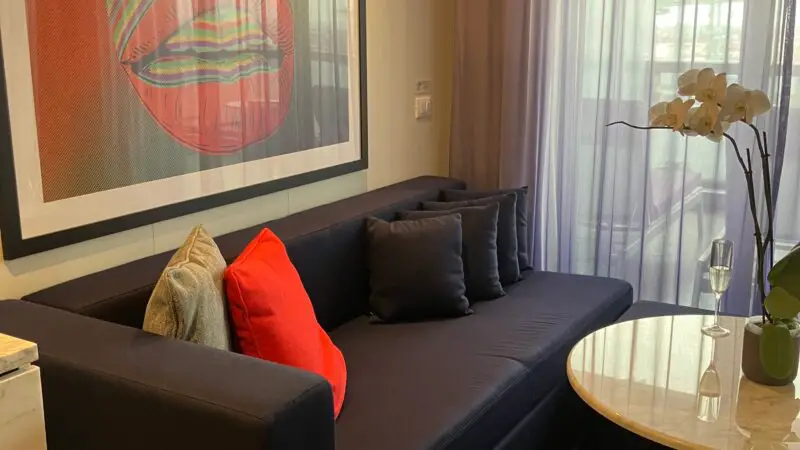 Virgin Voyages cruise ship cabin with a modern sofa, colorful artwork of lips, a round table, and balcony access with sheer curtains.
