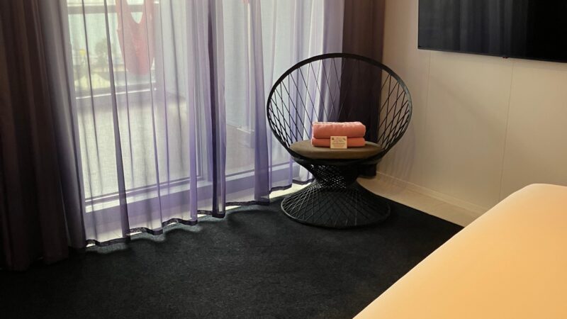 Cruise ship cabin with a bed, folded towels, a chair, and a TV. Sheer curtains cover a window leading to a balcony.