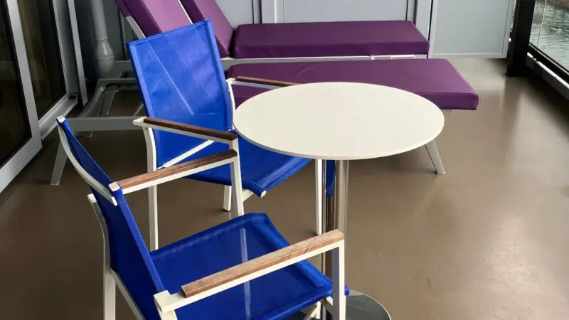 Balcony of a Virgin Voyages cruise ship cabin featuring a blue chair, a small round table, and a purple lounge chair.