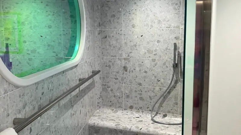 Modern bathroom in a Virgin Voyages cruise ship cabin, featuring terrazzo tiles, a rain shower head, a bench, and a towel rail beneath a decorative mirror.