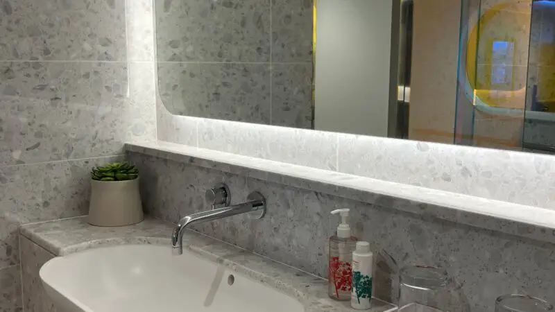 Luxurious Virgin Voyages cruise ship cabin bathroom with a sleek white sink, modern chrome faucet, large mirror, folded towels, toiletries, and speckled stone surfaces.