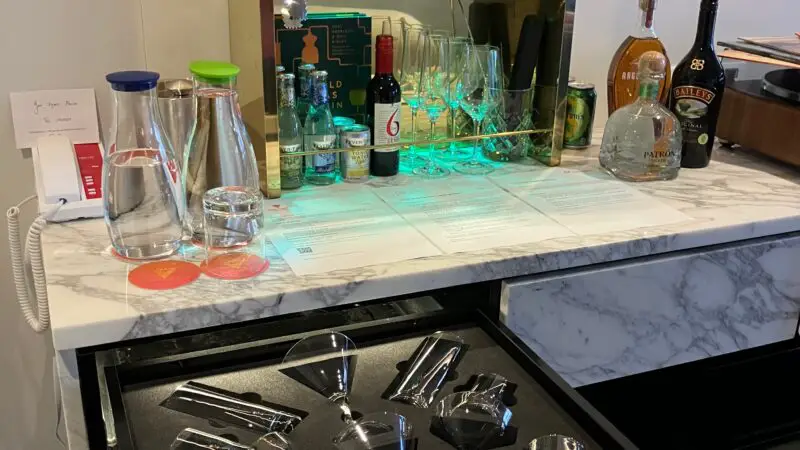Virgin Voyages cruise ship cabin minibar with assorted bottles, glassware, and open drawer showcasing cocktail glasses.