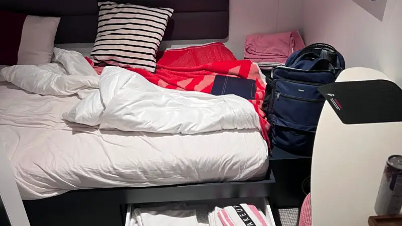 Virgin Voyages cruise ship cabin with unmade bed, striped pillows, open drawer containing towels, and a closed navy backpack next to a small desk.