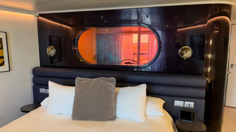 Virgin Voyages cruise ship cabin with a bed, dark headboard, round window, and modern decor.