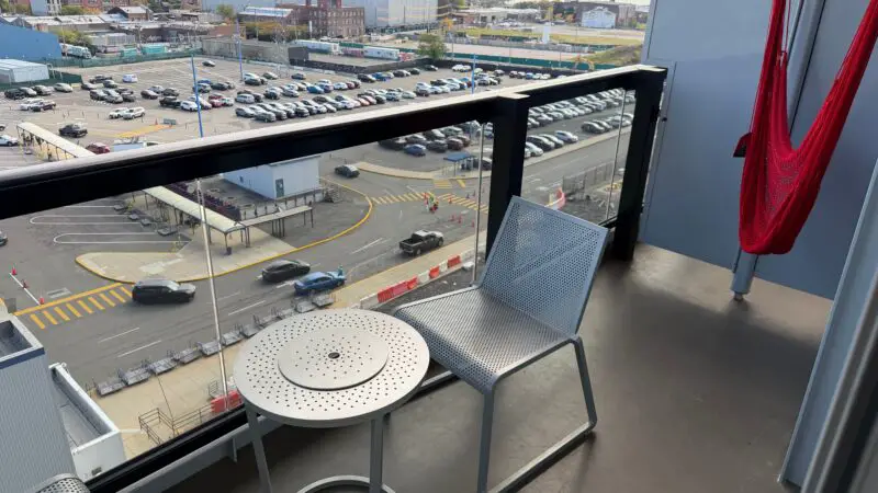 Balcony of a Virgin Voyages cruise ship cabin with two metal chairs, a small table, and a red hammock, overlooking a parking lot and city view.