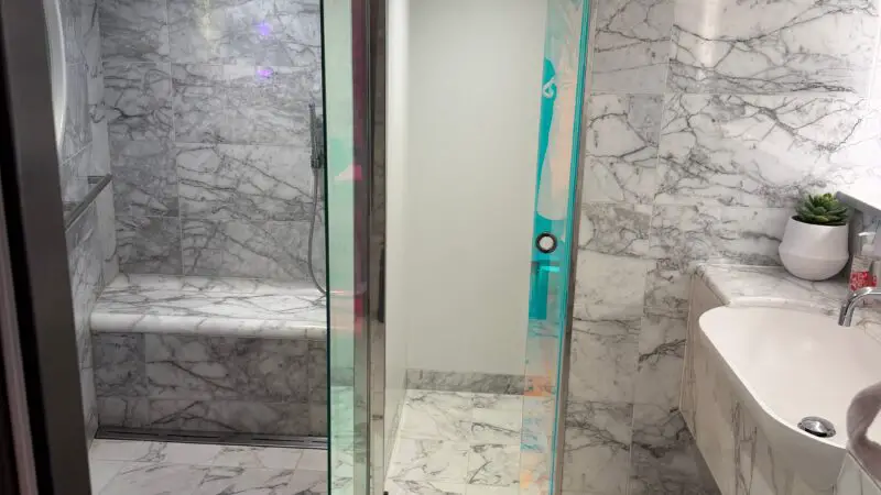 Luxurious Virgin Voyages cruise ship cabin bathroom featuring marble walls and floors, a glass shower enclosure, a sleek white sink, towel, and small plant for decor.