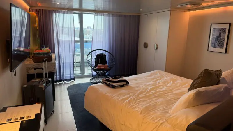 Virgin Voyages cruise ship cabin with a white bed, wall-mounted TV, and sliding glass doors leading to a balcony.