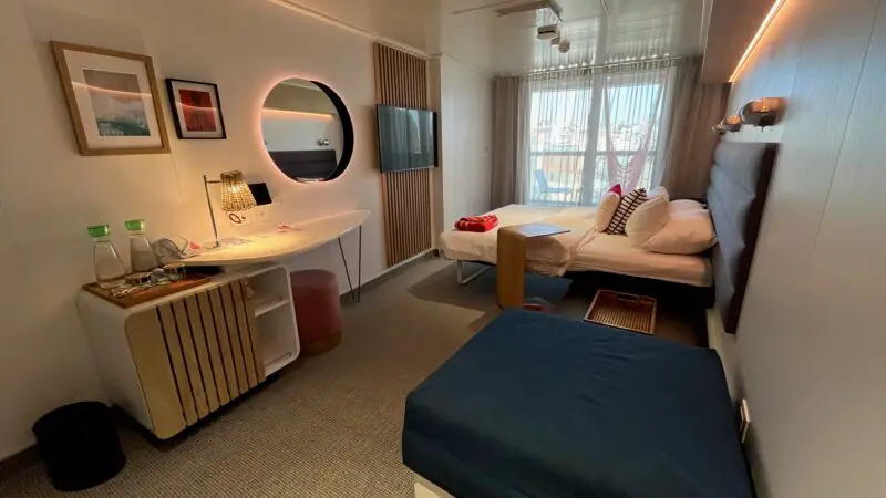 A modern Virgin Voyages cruise ship cabin with a bed, sofa, wall-mounted TV, and balcony window. A desk with a mirror and art decorates the space.