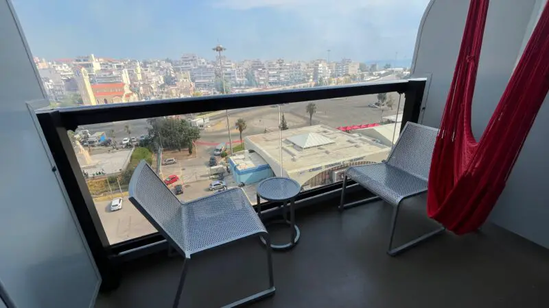 Virgin Voyages cruise ship stateroom balcony with two metal chairs, a small table, and a red hammock, overlooking a cityscape.