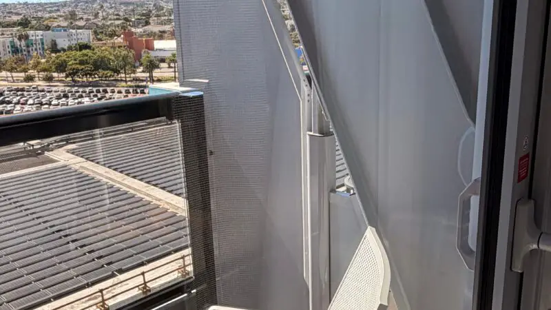 Cruise ship cabin balcony with a white chair and round table, overlooking a cityscape and parking lot.