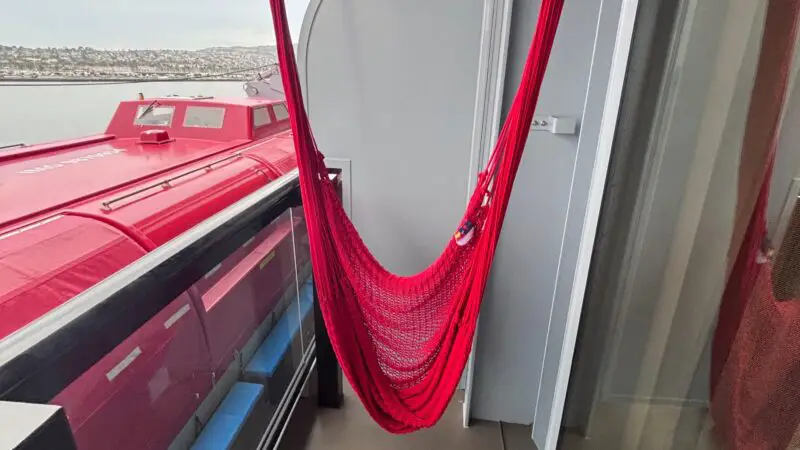 Cruise ship balcony with a vibrant red hammock, ocean and distant shoreline visible in the background.