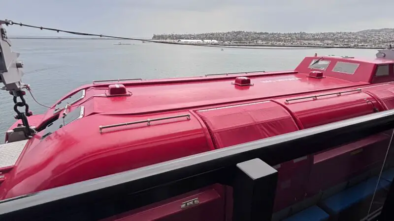 Balcony view from a Virgin Voyages cruise ship cabin, overlooking the ocean with a bright red lifeboat visible below the railing.