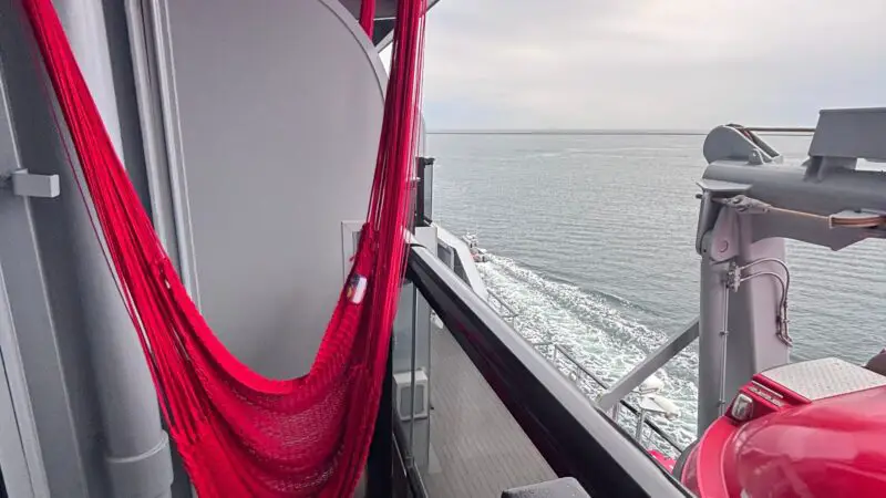 Balcony of a Virgin Voyages cruise ship cabin featuring a red hammock, small table, and ocean view.