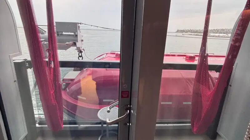 View from a Virgin Voyages cruise ship cabin showing a balcony with red hammocks, a small table, and a partial ocean view.