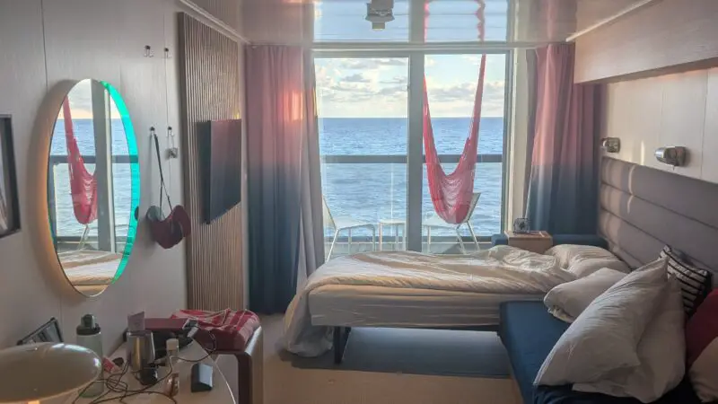 A Virgin Voyages cruise ship cabin with a bed, sofa, and balcony overlooking the ocean. A red hammock hangs on the balcony with sunlight filtering through curtains.