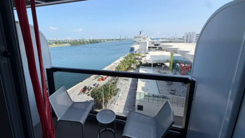 View from a Virgin Voyages cruise ship cabin balcony with two chairs, a table, and a hammock, overlooking a port and the sea.