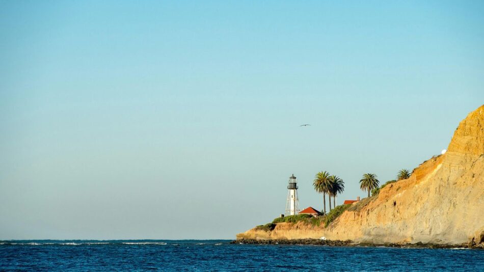 A distant lighthouse stands on a rocky cliffside with palm trees, overlooking a calm blue sea under a clear sky.